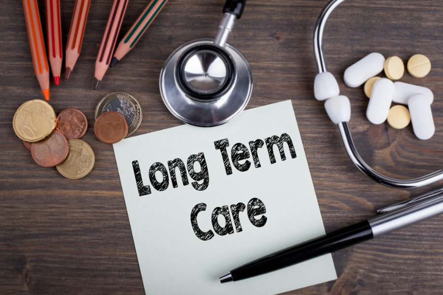 Coins, a colored pencils, a stethoscope, pills, and a sheet of paper with the words "Long Term Care" written on it all on a desktop.