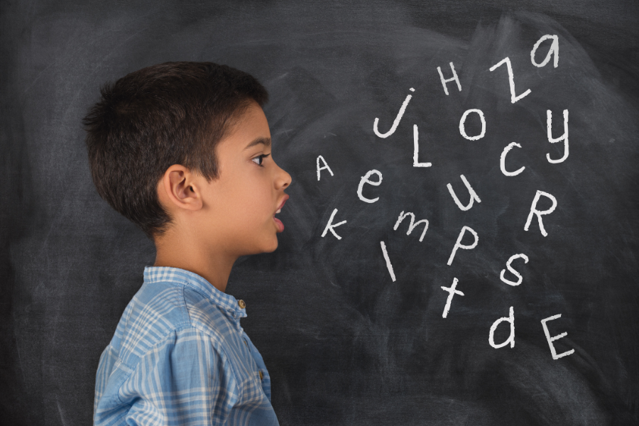 A young boy in front of a chalkboard. There are written letters on the chalkboard that appear to be coming from his mouth.
