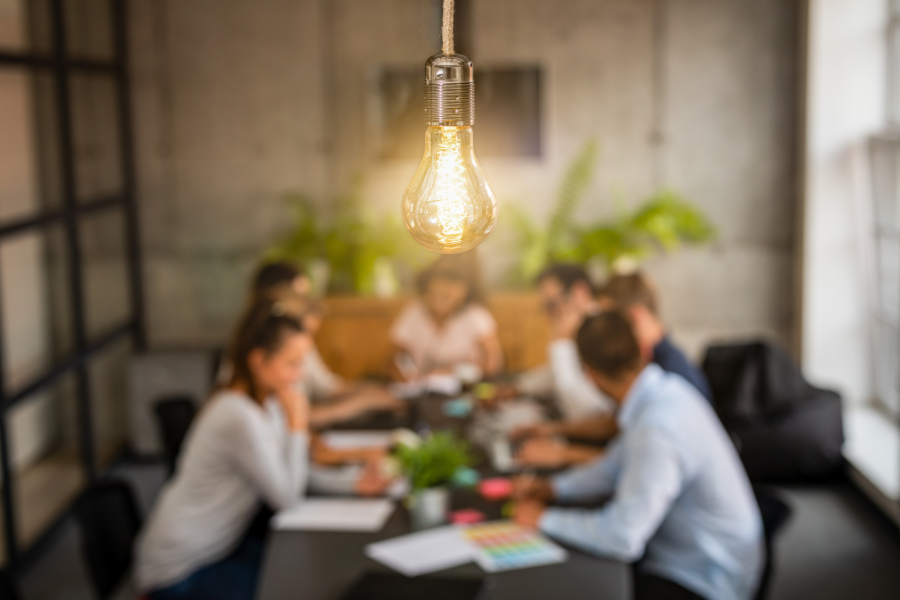 A bright lightbulb hangs down, with a group having a meeting around a table blurred out in the background.