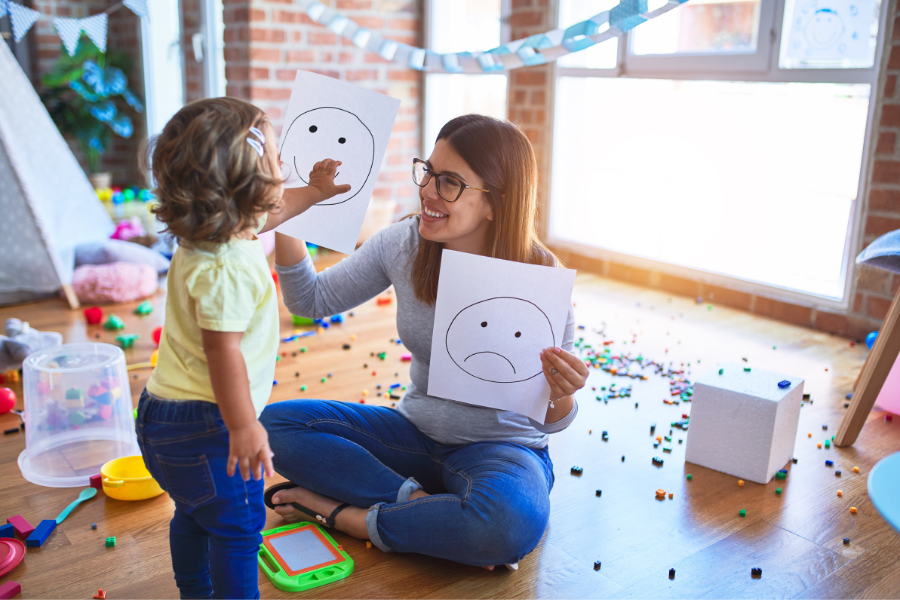A therapist holds up two drawings: a frowning face and a smiling face. A young child points to the picture of a smiling face.